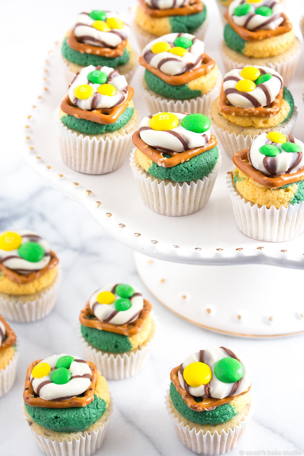 a white cake plate filled with go big or go home team cupcakes: mini cupcakes with yellow and green frosting and a team pretzel hug on top.
