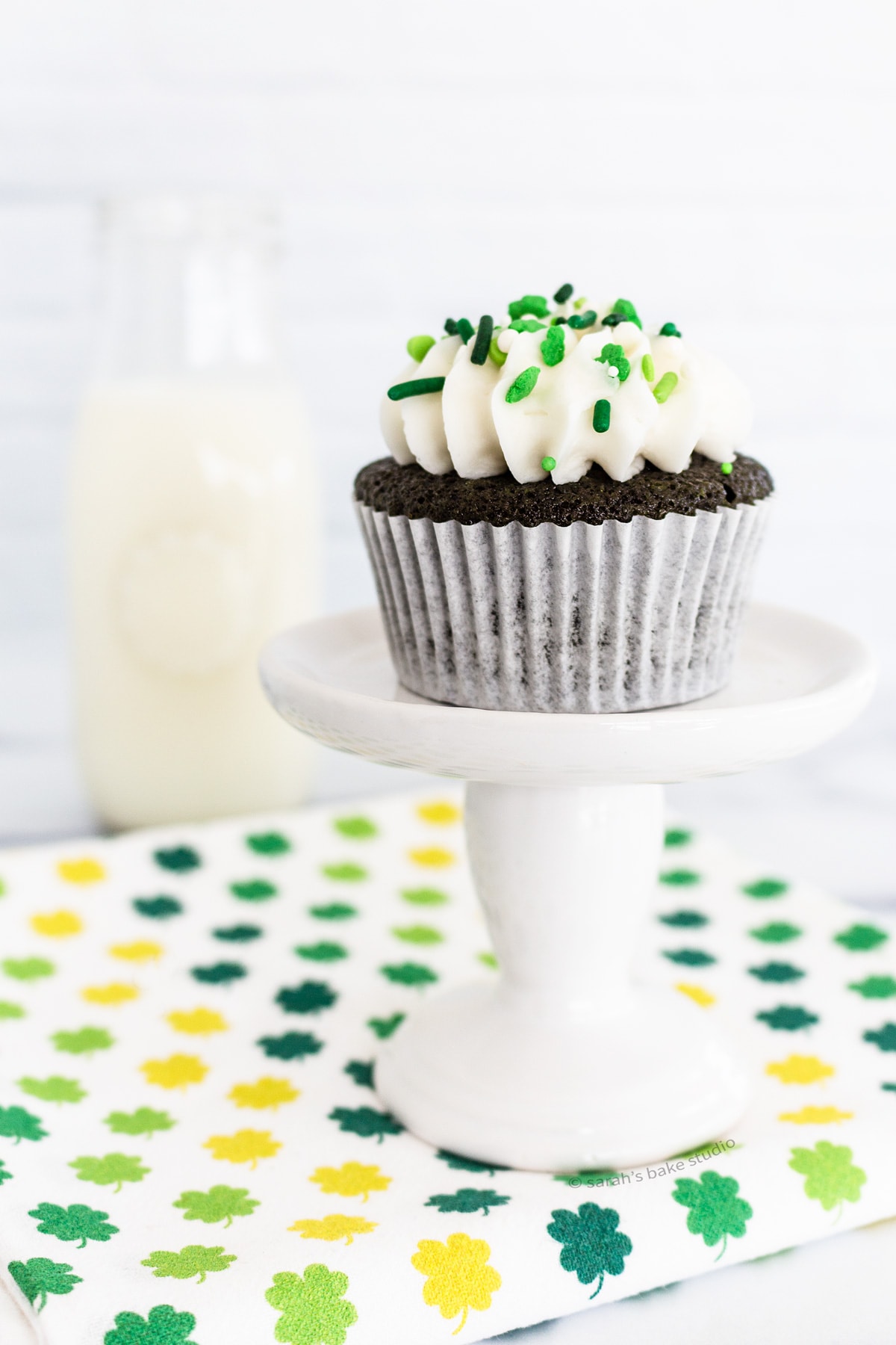 A bottle of milk and a cupcake stand with a Green Velvet Cupcake on it.