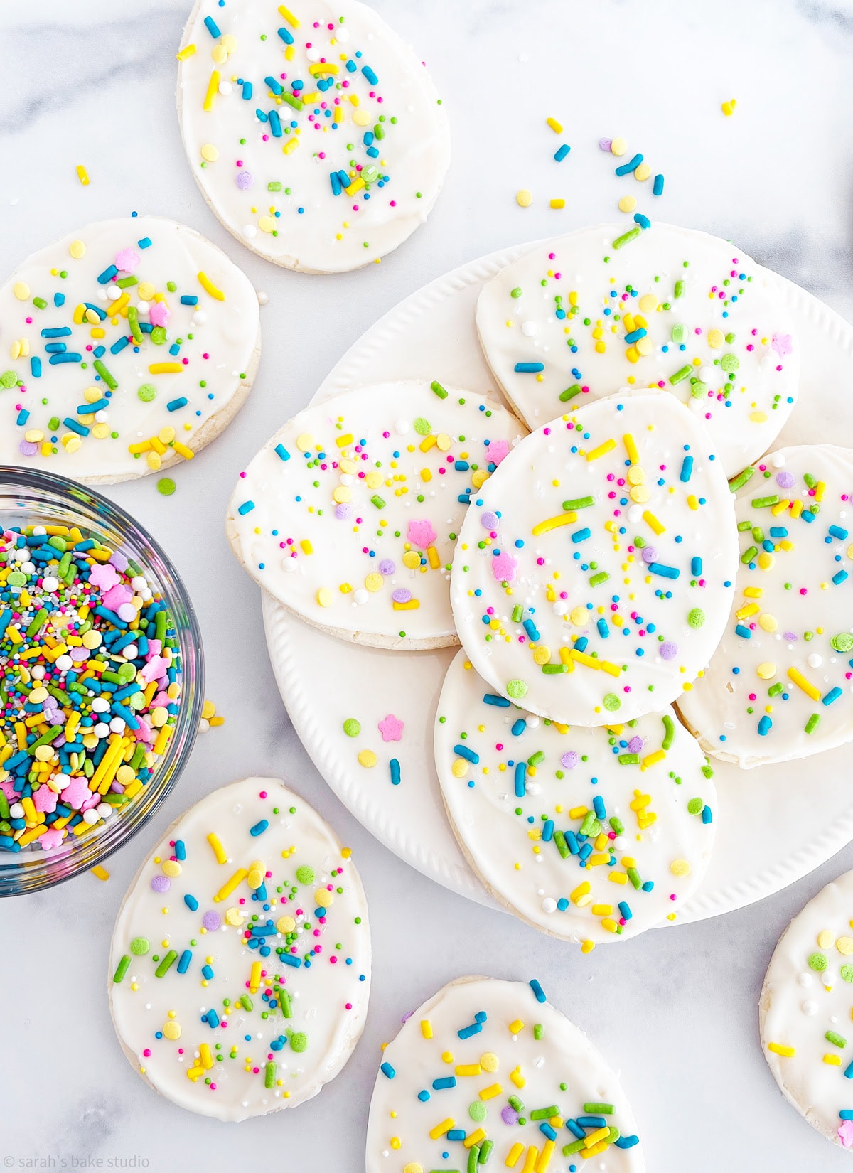 A plate stacked with White Chocolate Easter Egg Sugar cookies and a bowl full of spring sprinkles.