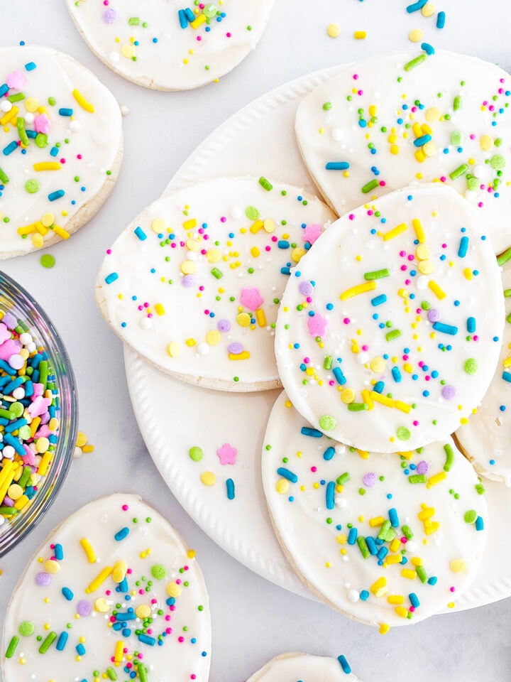 A plate stacked with White Chocolate Easter Egg Sugar Cookies and a bowl of spring sprinkles.